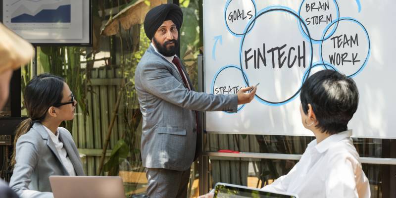 Image of a person pointing at the word FINTECH mentioned on a white board and two women taking part in the discussion.
