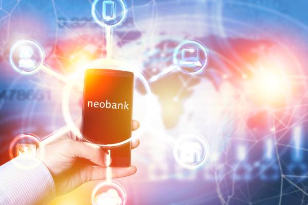 A persons hand holding a mobile with the word neobank displayed on the screen and related icons shown as the background.