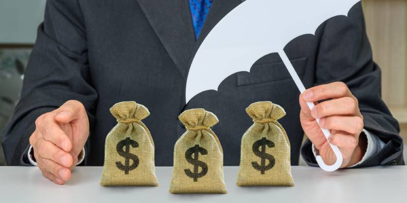 An investor holds an umbrella protecting dollar bags, depicting a plan to avoid risk and protect asset or business.