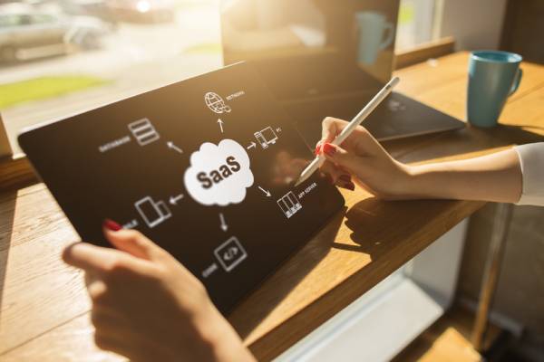 A women holding a pen and a tablet showing the term SAAS in the middle with the SAAS icons around it.